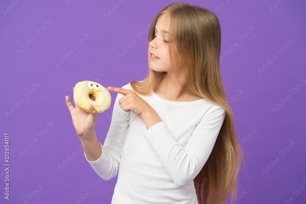 Girl holding fluffy doughnut. Glazed bagel with eyes, creative dessert. Kid eating icing out off doughnut, sweet tooth heaven. Child with long hair wearing white jumper isolated on purple background