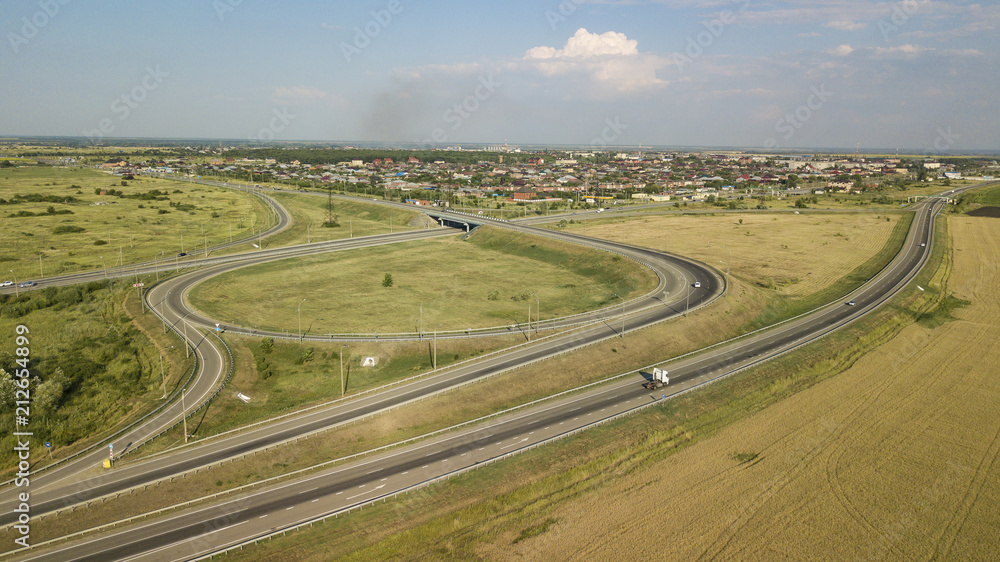 Summer aerial drone photo of transport junction, traffic cross road junction day view from above with green tree circle road, near is a wheat field. Top down view of traffic jam.