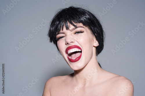 Woman with attractive red lips looks at camera. Girl on scandalous shouting face posing with naked shoulders. Lady in black wig with make up on grey background. Scandalous lady concept photo
