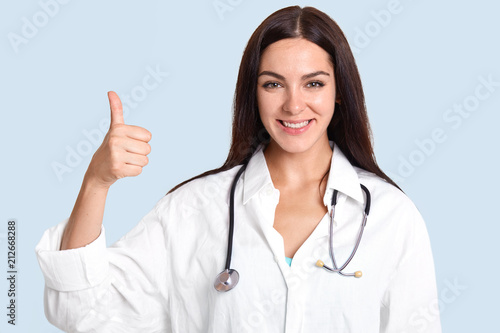 Horizontal shot of pretty talented female doctor with satisfied expression  raises thumb  shows like gesture  approves something  likes her job and helping people  isolated over blue background