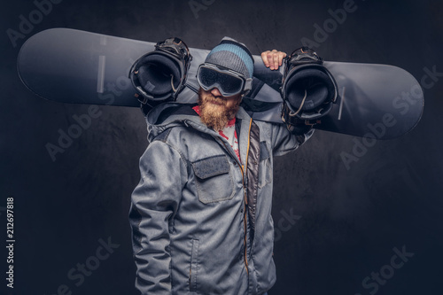 Brutal redhead snowboarder with a full beard in a winter hat and protective glasses dressed in a snowboarding coat holds a snowboard on his shoulder in a studio. Isolated on a gray background.