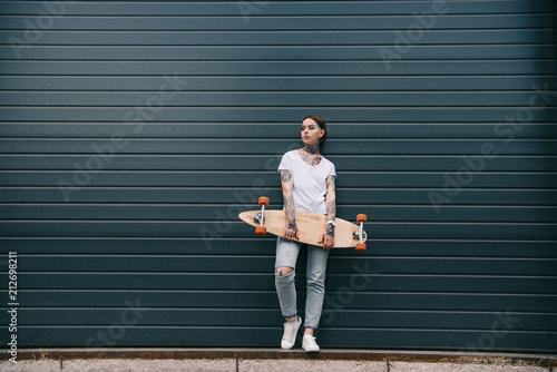 distant view of young woman with tattoos holding skateboard against black wall