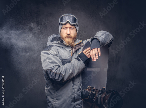 Brutal redhead snowboarder with a full beard in a winter hat and protective glasses dressed in a snowboarding coat posing with snowboard at a studio, looking away. Isolated on gray background.