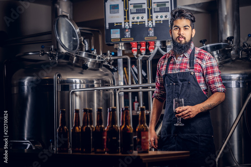 Stylish full bearded Indian man in a fleece shirt and apron holds a glass of beer, standing behind the counter in the brewery.
