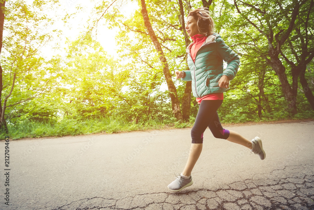 Young fitness blonde woman in headphones running at morning caucasian forest trail in sun light Young fitness blonde woman in headphones running at morning caucasian forest trail in sun light