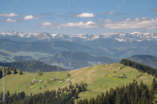 Scenic shot of rural area at mountains taken at sunny spring day. Mountains covered with forest and meadows, snowy mountains and blue sky with clouds. Concept of clear environment. Natural background