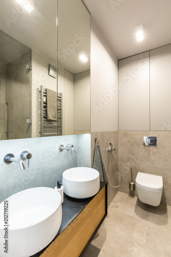 Mirror above white washbasins in beige bathroom interior with light and toilet. Real photo