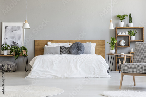 Patterned armchair in grey bedroom interior with poster on table with plants next to bed. Real photo