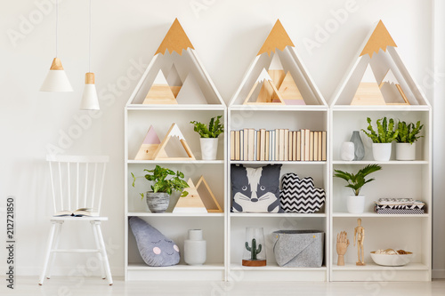 Lamps above white chair next to shelves with wooden triangles and plants in bright interior. Real photo