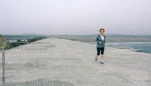 Senior sportswoman with headphones running by sea pier