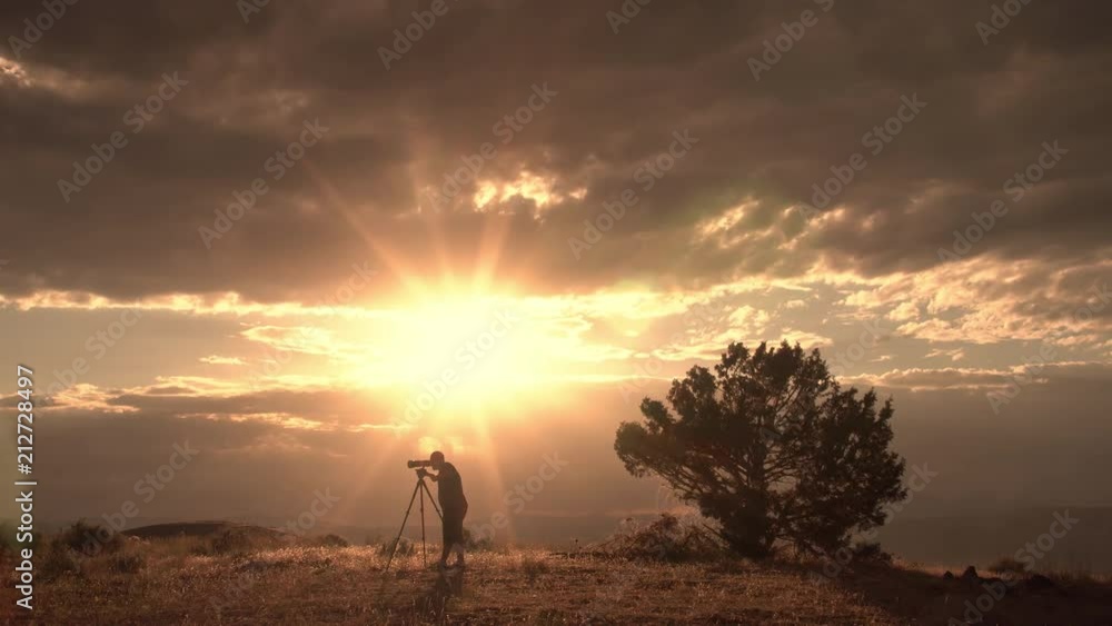 Photographer looking through sunset landscape on hill top next to lone tree as sun rays shine.