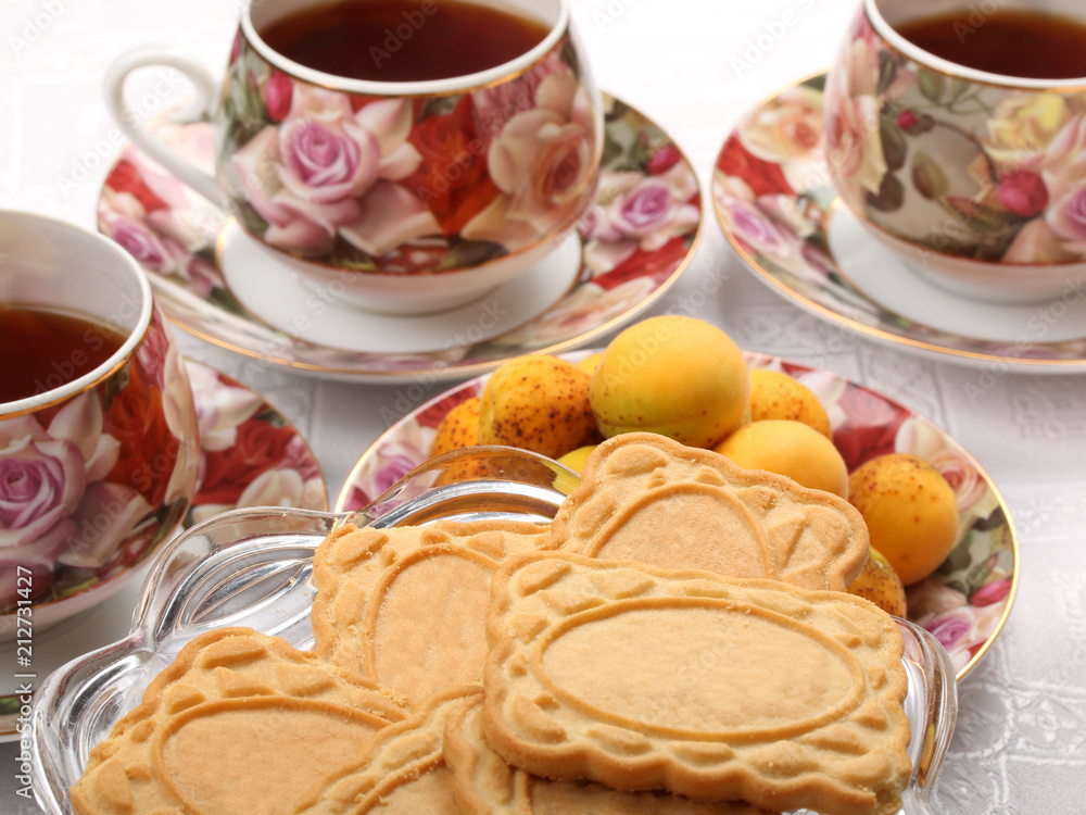 Porcelain teacups with hot tea and with floral pattern, biscuits and plate with wild apricots on a table Porcelain teacups with hot tea and with floral pattern, biscuits and plate with wild apricots on a table