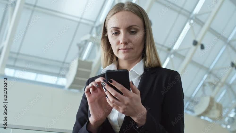 Attractive young businesswoman with blonde hair standing at glass balustrade in large modern office building and typing on smartphone Attractive young businesswoman with blonde hair standing at glass balustrade in large modern office building and typing on smartphone