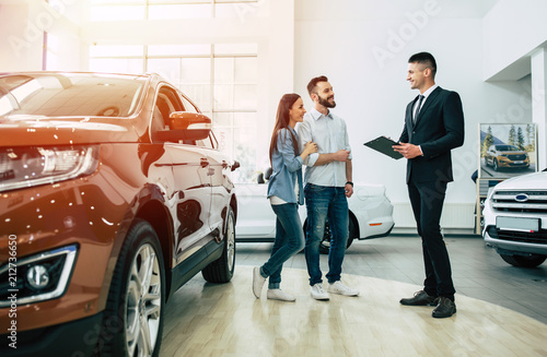 Young modern happy couple buying a new car in city dealership and talking with salesman