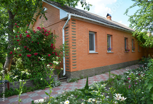 Cozy brick house with asbestos roof and red clibing roses, beautiful flower bed. photo