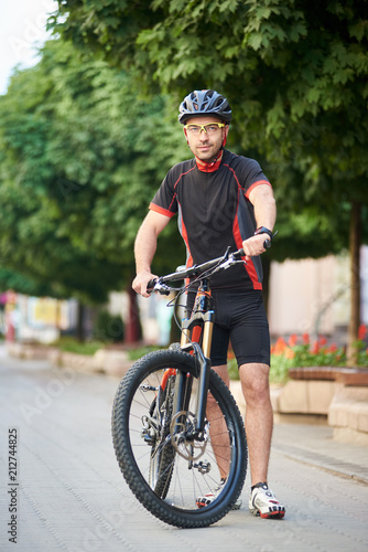 Young male bicyclist in cycling clothes and protective helmet standing near bike in empty city street. Sportsman shooting for sport ad clothing campaign. Concept of healthy lifestyle