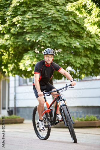 Young male bicyclist in cycling sportswear and protective helmet, looking at camera, riding bike down empty city center alley surrounded by green trees. Sportsman training outdoors improving skills.