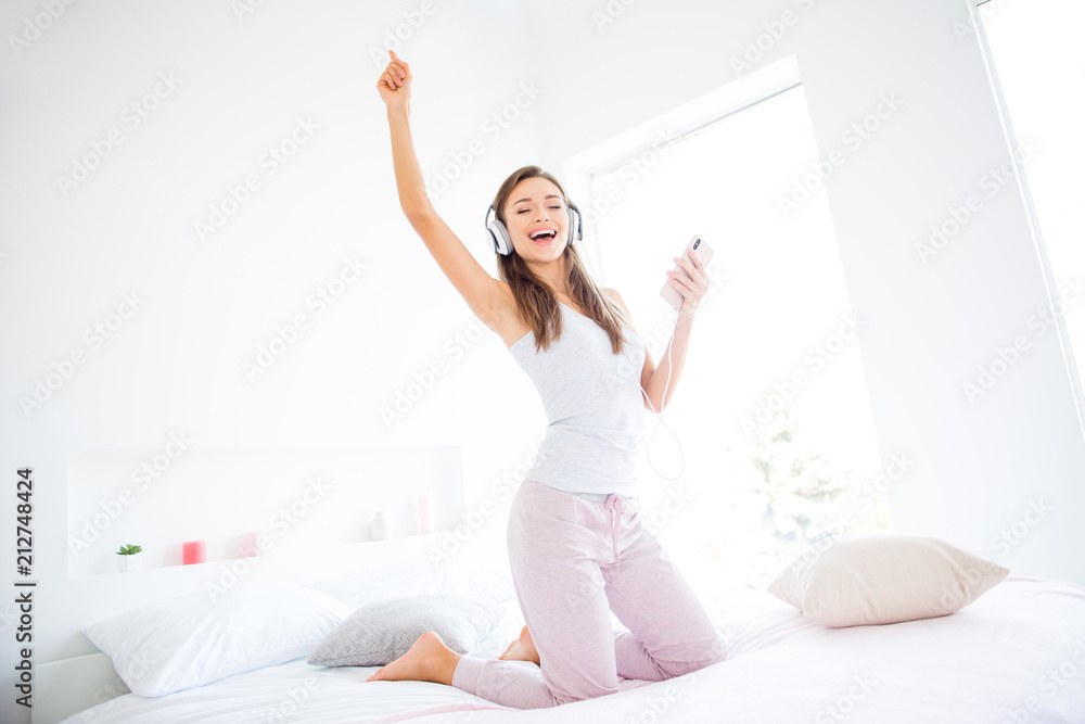 Portrait of cheerful positive girl dancing on bed using smart phone and headset listening favorite music enjoying morning time Portrait of cheerful positive girl dancing on bed using smart phone and headset listening favorite music enjoying morning time