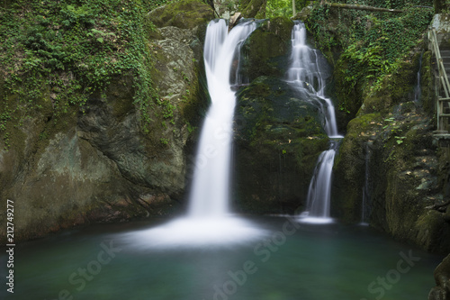 Fototapeta Naklejka Na Ścianę i Meble -  Water flows down a gentle, rocky stream in the mountains surrounded by lush green foliage in this long exposure scenic landscape.