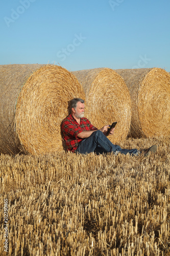 Farmer or agronomist in wheat field after harvest examining bale, rolled straw, using tablet