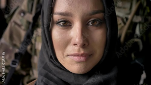 Close portrait of young muslim woman in hijab crying and looking at camera, armed soldier with weapon standing behind woman, war photo