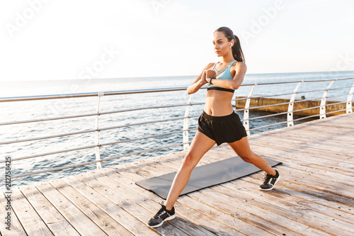 Image of energetic brunette sportswoman 20s in tracksuit doing sports, and stretching body on boardwalk near seaside with bluetooth earbud