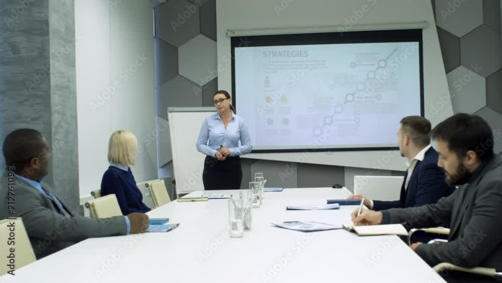 Businesswoman standing at whiteboard and explaining presentation to diverse team of coworkers sitting at meeting table Businesswoman standing at whiteboard and explaining presentation to diverse team of coworkers sitting at meeting table