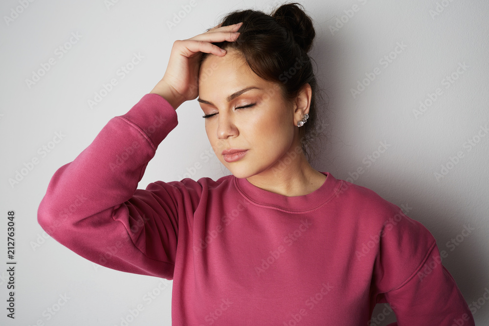 Beautiful young woman with closed eyes wearing pink hoody and holding hand on head over empty white background.Model with light nude make-up, studio background, copy paste text space. Beautiful young woman with closed eyes wearing pink hoody and holding hand on head over empty white background.Model with light nude make-up, studio background, copy paste text space.