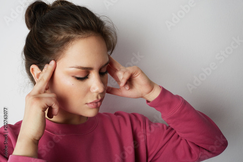 Handsome woman in pink hoody with a severe headache with the hands in the head over white empty wall on background. Beauty and healthcare concept