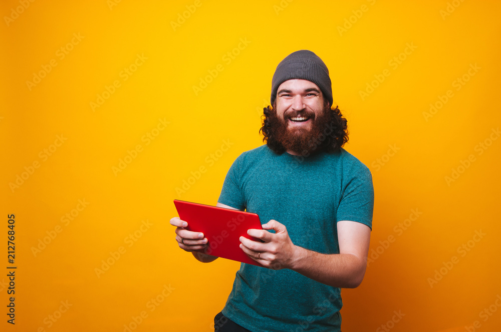 Playful bearded man looking at the camera and playing on his red tablet. Man wearing fall hat fur cap on yellow background. Playful bearded man looking at the camera and playing on his red tablet. Man wearing fall hat fur cap on yellow background.
