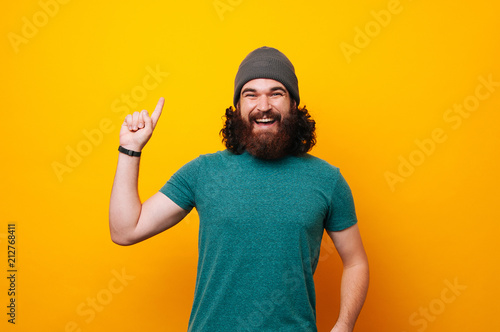 Laughing bearded man wearing fur cap hat, looking at the camera and poining upwards on yellow background. photo