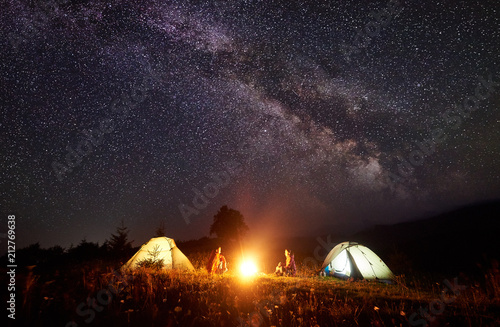 Night camping in mountains. Bright campfire burning between two backpackers, man and woman sitting opposite each other in front of illuminated tents under amazing dark blue starry sky and Milky way