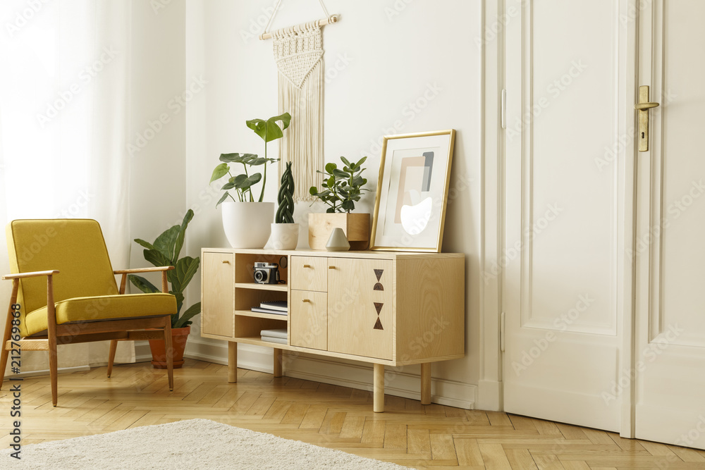 Fototapeta premium Yellow wooden armchair next to cupboard with plants and poster in retro flat interior. Real photo