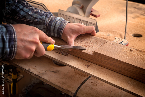 Close up of young man builder wearing in a plaid shirt treating a wooden product with a chisel in the workshop, close-up