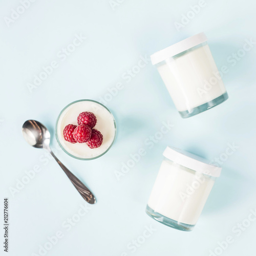 Open jar with homemade yogurt and raspberries next to closed jars on a blue background. Top view, flat lay