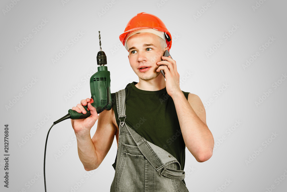 Young working guy in hard hat holds drill and calls by phone isolated on a gray background Young working guy in hard hat holds drill and calls by phone isolated on a gray background