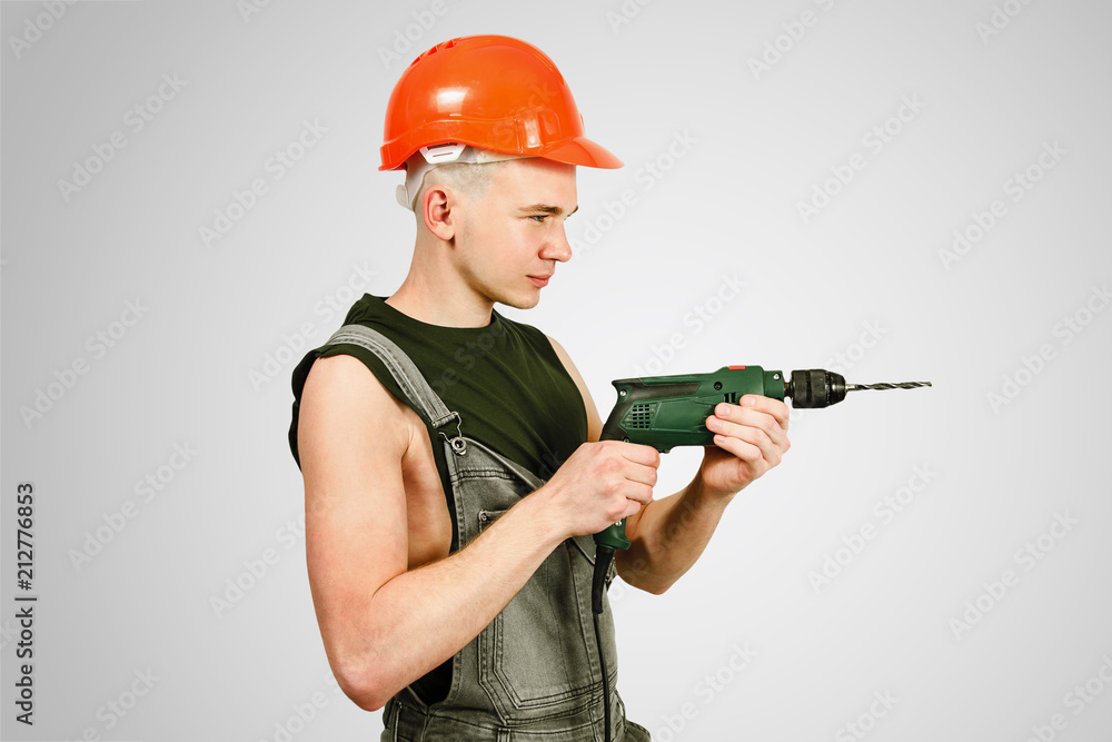 Young working guy in hard hat holds drill isolated on a gray background Young working guy in hard hat holds drill isolated on a gray background
