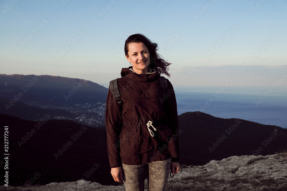 A girl in a jacket and with a backpack is standing on top of a mountain overlooking the sea in the rays of the setting sun.