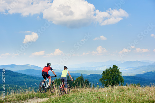 Rear view of active couple bikers in professional sportswear riding cross country bicycles down on the mountain road under bright blue sky with clouds on summer day in the Carpathian mountains.