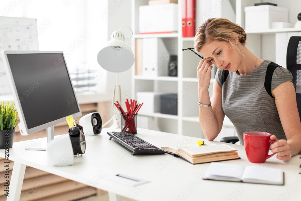 A young girl sits at a table in the office and holds a red cup and glasses in her hands. Before the girl lies an open book. A young girl sits at a table in the office and holds a red cup and glasses in her hands. Before the girl lies an open book.
