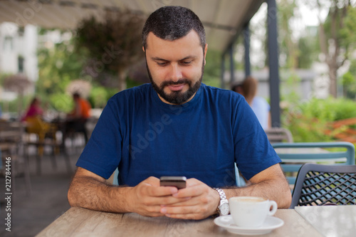 Smiling young man texting message on a smart phone in cafe