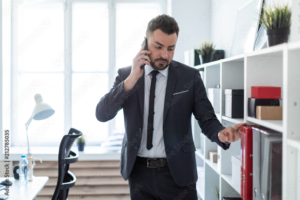 The man is standing near the shelves in the office, talking on the phone and putting his hand on the folder. The man is standing near the shelves in the office, talking on the phone and putting his hand on the folder.