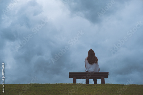 A woman sitting alone on a wooden bench in the park with cloudy and gloomy sky background