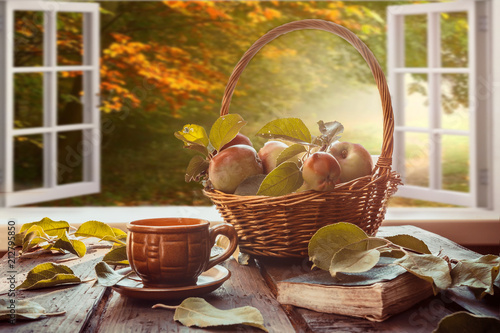 Apples in a basket, a plaid book, a cup on the table near a window overlooking the autumn landscape, autumn still life, the concept of coziness in a rural house during harvesting