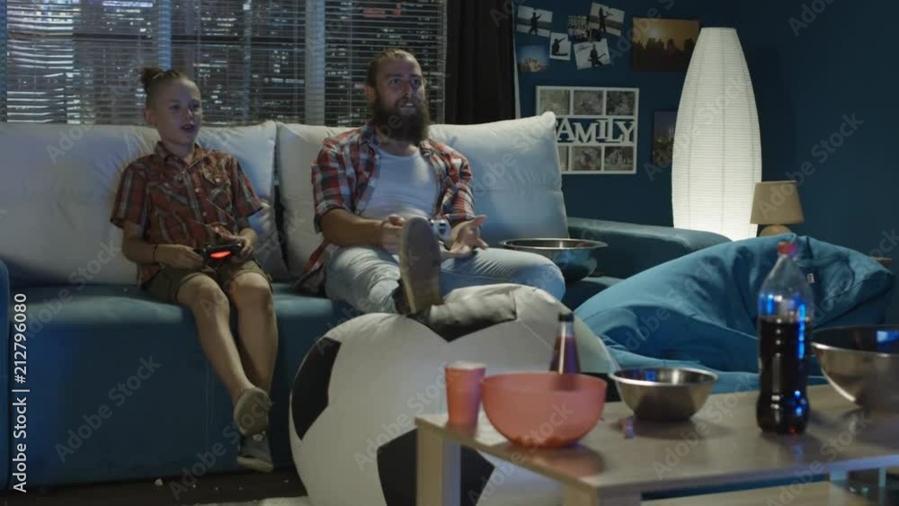 Teen boy defeating father in videogame playing and mocking while having fun together on sofa at home Teen boy defeating father in videogame playing and mocking while having fun together on sofa at home