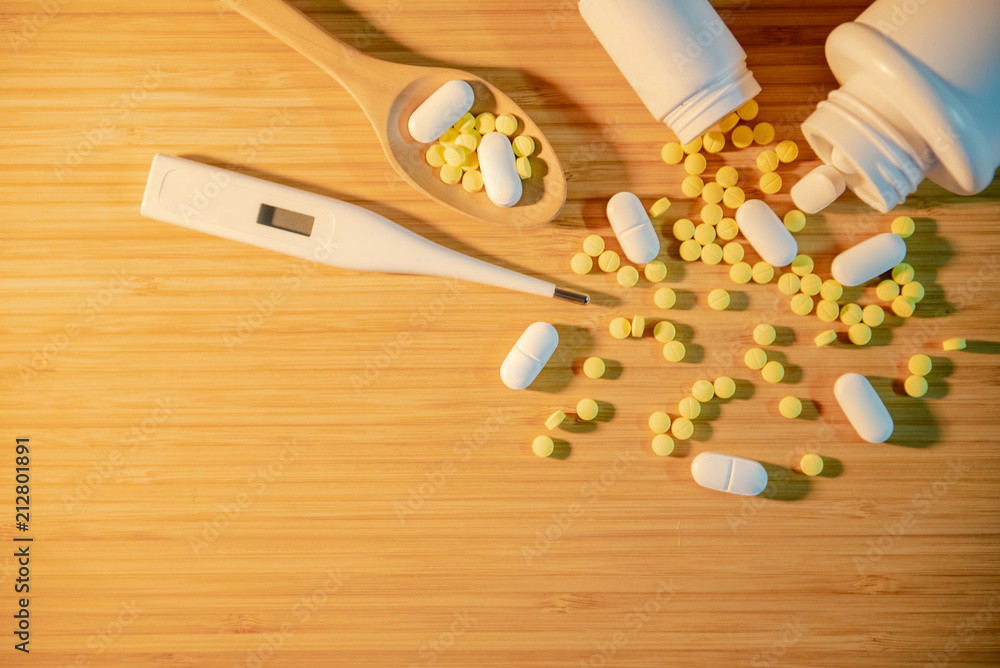 Pills spilling out of pill bottles with digital thermometer and pharmaceutical spoon on wooden table. Prescription medicine and medical treatment background concepts Pills spilling out of pill bottles with digital thermometer and pharmaceutical spoon on wooden table. Prescription medicine and medical treatment background concepts