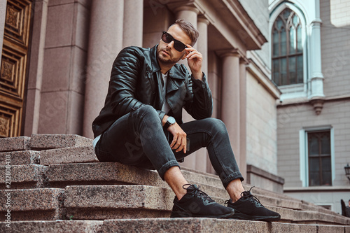 Fashionable guy dressed in a black jacket and jeans holds the smartphone sitting on steps against an old building in Europe. photo