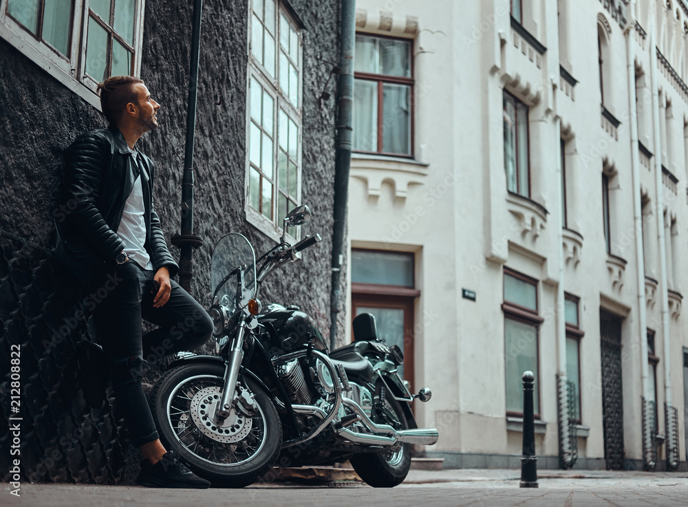 Fashionable biker dressed in a black leather jacket and jeans leaning on a wall near his retro motorcycle on old Europe street.