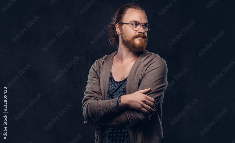 Portrait of a redhead hipster male dressed in casual clothes with glasses and full beard, standing with crossed arms in a studio. Isolated on the dark background.