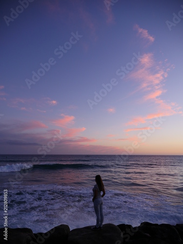 woman from behind standing on rock at the ocean shore in front a breaking wave during sunset with pink clouds